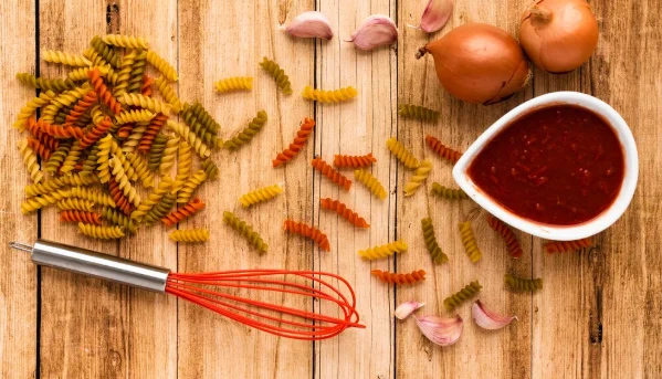 Raw pasta and ingredient with whisk on wooden table