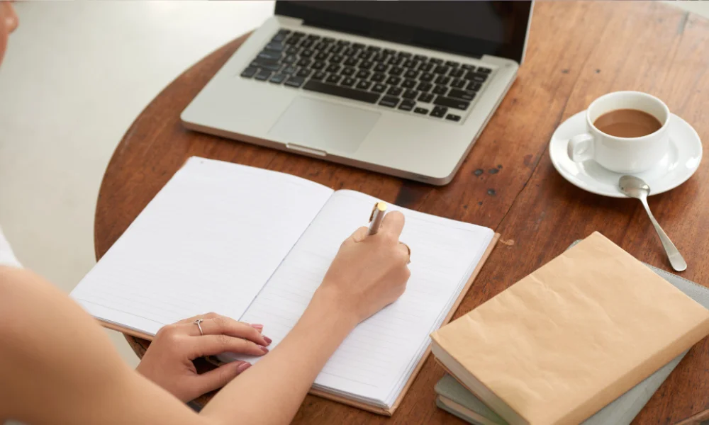 Close-up of hands writing on notebook with laptop open to planning workspace.