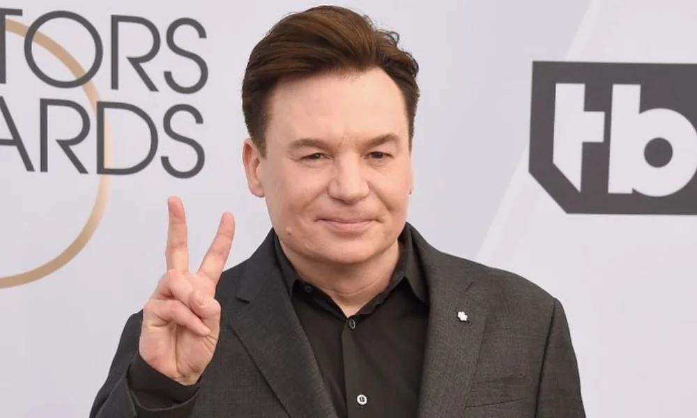 person in a dark suit flashes a peace sign-mike myers net worth