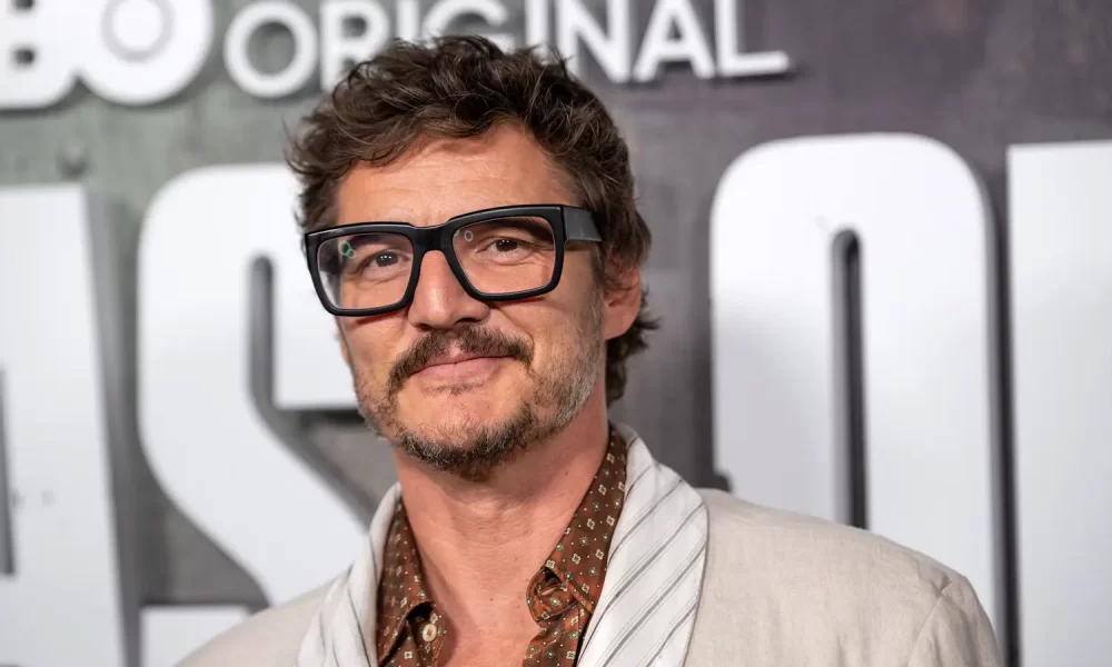Pedro Pascal wearing glasses and a light-colored jacket at a red carpet event.