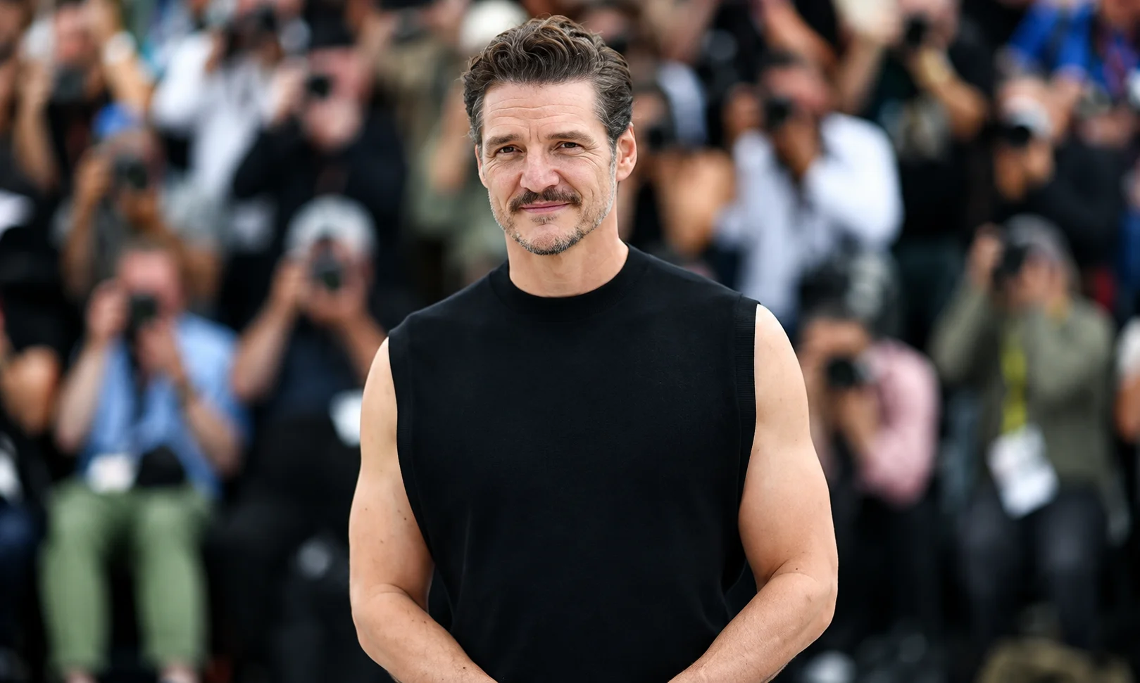 Pedro Pascal posing in a black sleeveless shirt at a red-carpet event with photographers in the background.