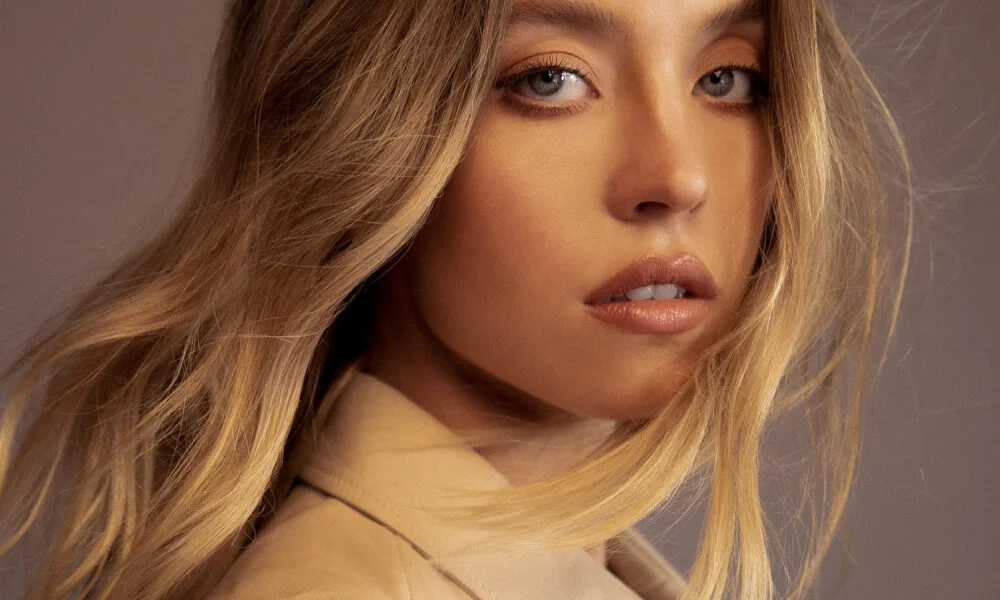 Sydney Sweeney in a close-up portrait with long blonde hair, wearing a beige outfit against a warm neutral background. 