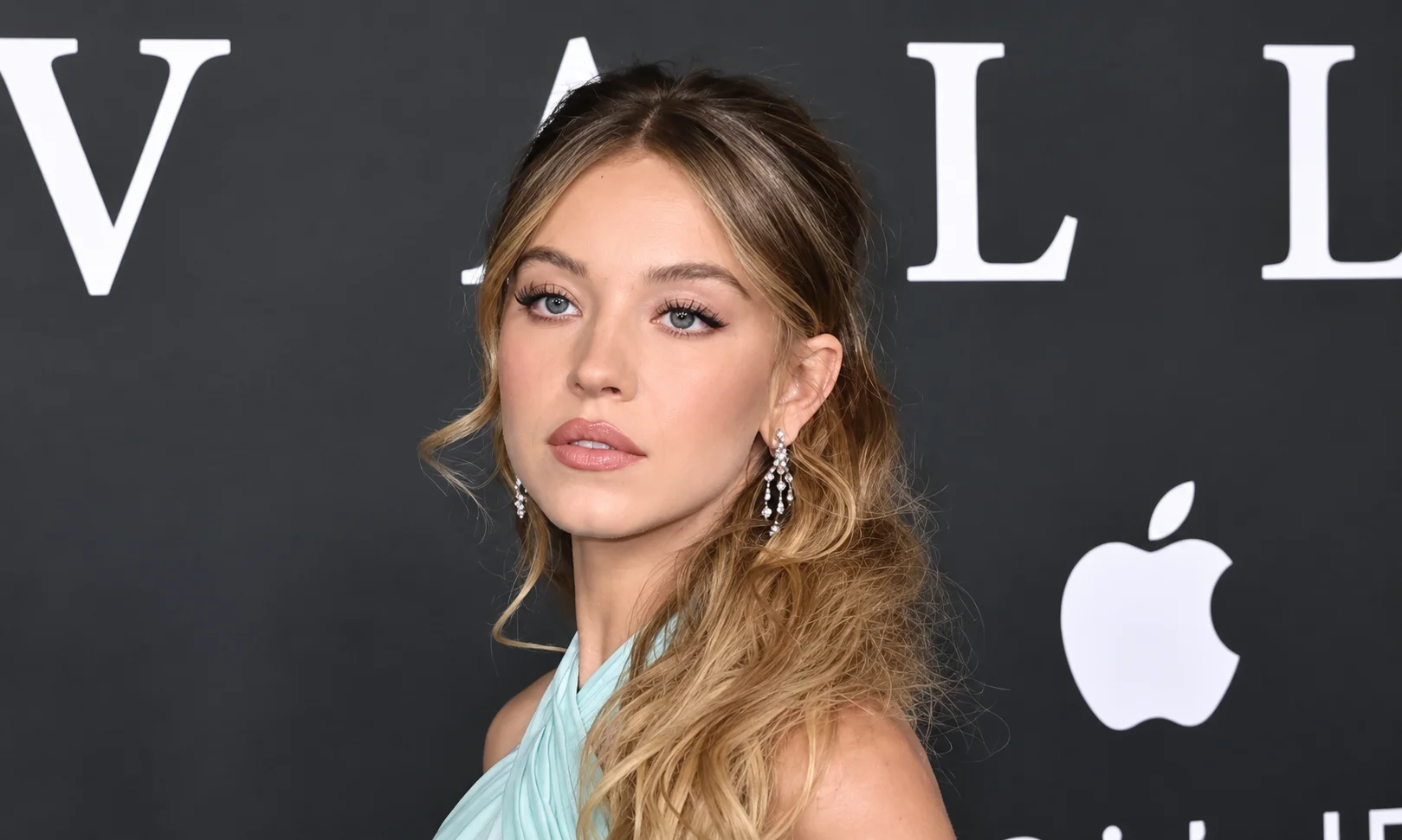 Sydney Sweeney wearing a light blue dress at a red-carpet event, posing in front of an Apple logo and event backdrop. 