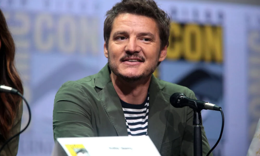 Pedro Pascal speaking into a microphone at a panel event while wearing a green jacket over a striped shirt.