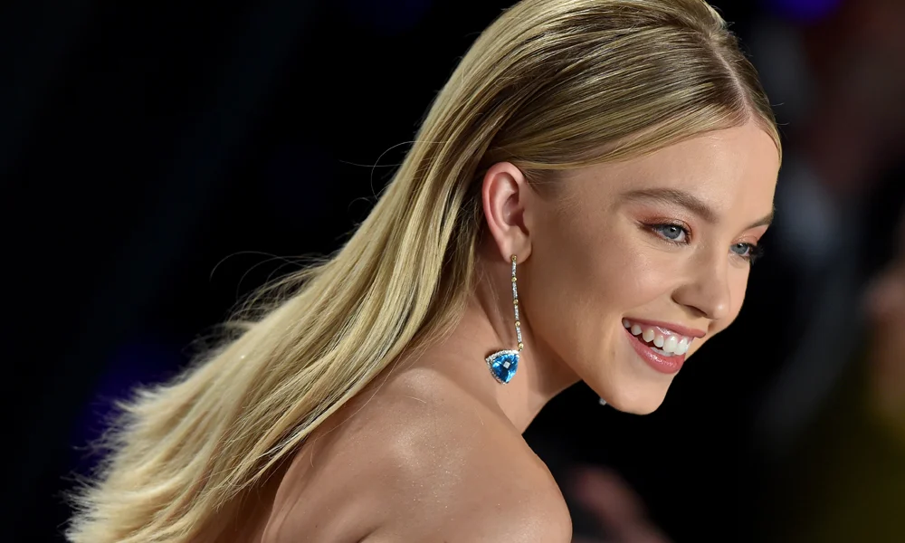Sydney Sweeney smiling at a red-carpet event, wearing a blue heart-shaped statement earring and posing against a dark background. 