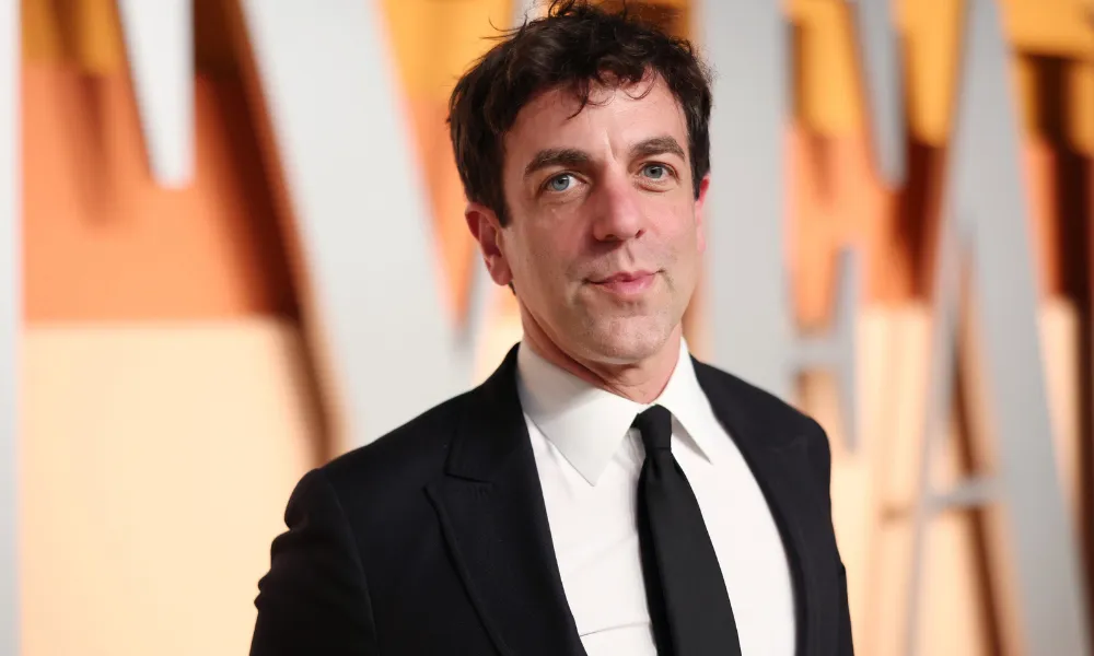 B.J. Novak Net Worth featured in a headshot of B.J. Novak wearing a formal black suit jacket, white dress shirt, and black tie at a red carpet event. 