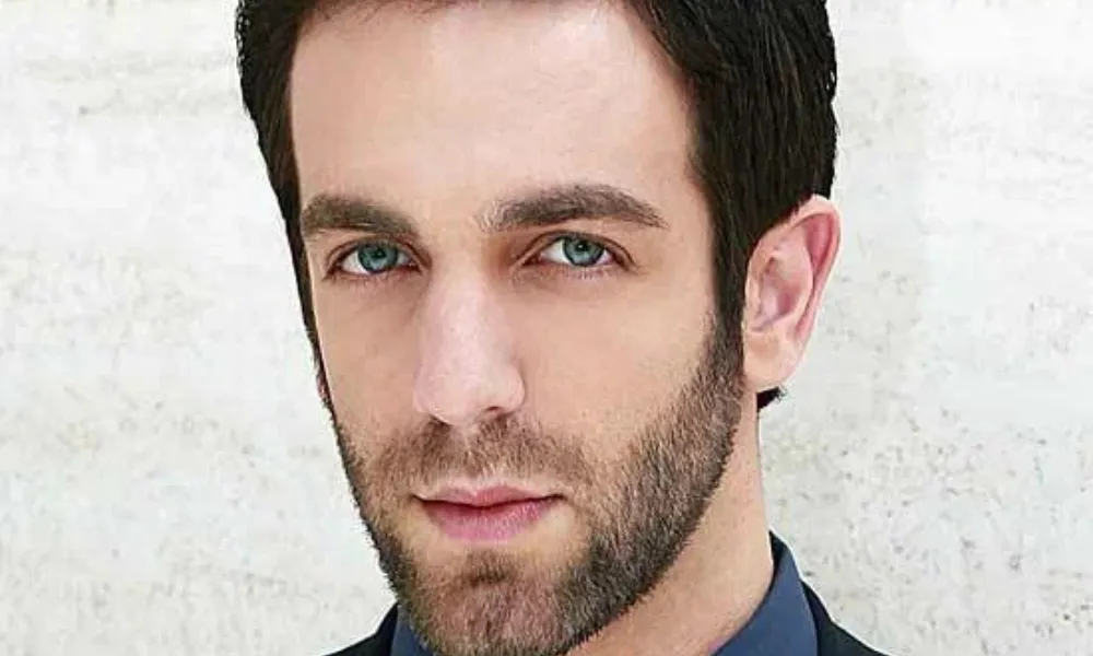 B.J. Novak Net Worth featured in a portrait of B.J. Novak with a trimmed beard wearing a navy blue dress shirt, dark tie, and black suit coat. 
