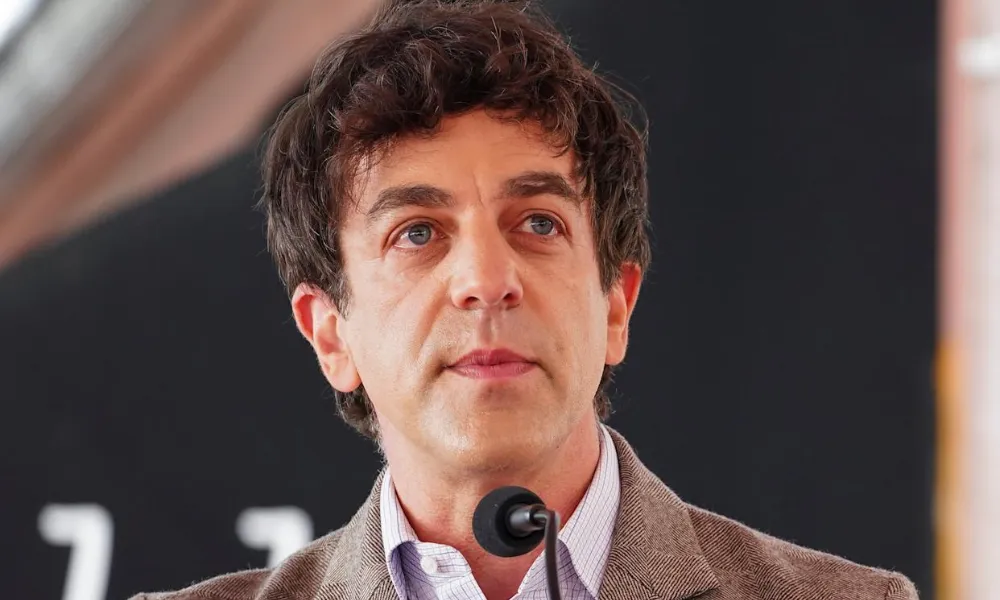 B.J. Novak Net Worth highlighted in a close-up photo of B.J. Novak speaking into a microphone while wearing a brown tweed jacket and collared shirt.