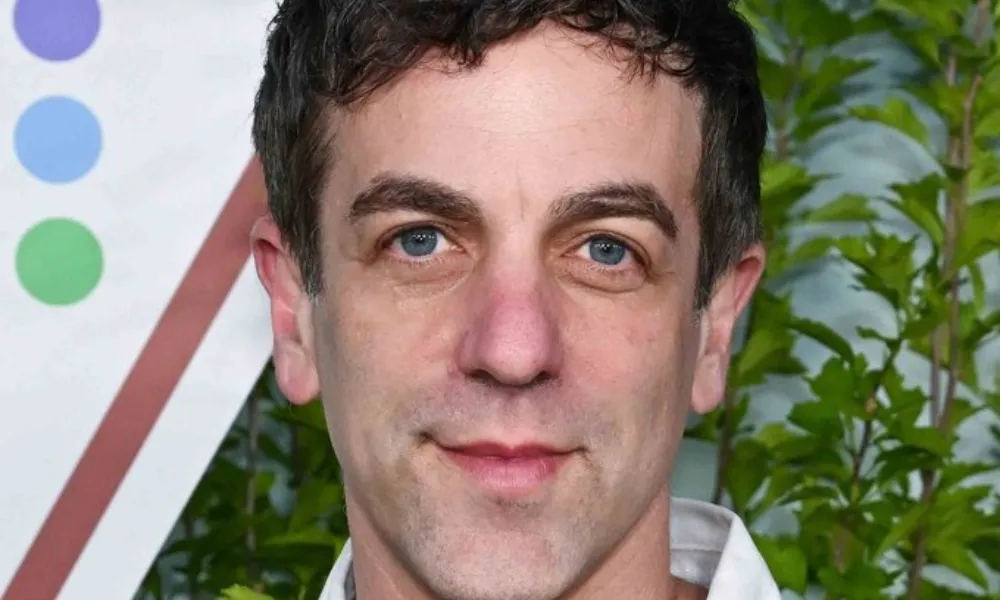 B.J. Novak Net Worth featured in a close-up portrait of B.J. Novak wearing a black polo sweater over a white open-collar shirt in front of leafy green foliage. 