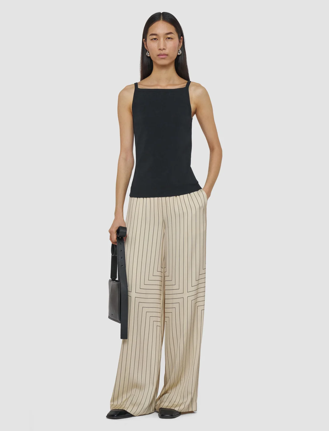 Model wearing a black square-neck tank top and beige wide-leg trousers with thin black geometric stripes, holding a black shoulder bag.