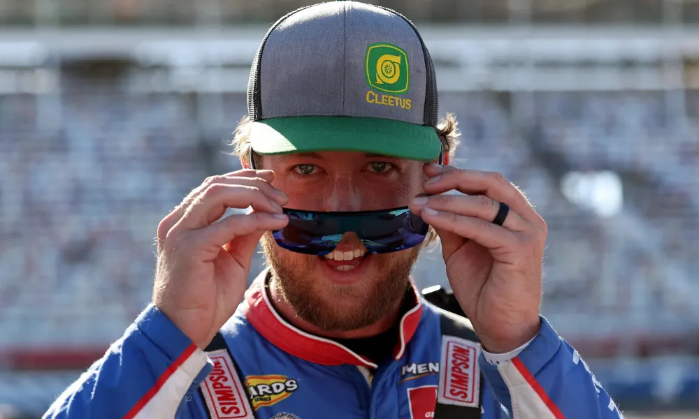 Cleetus McFarland Net Worth featured in a photo of Garrett Mitchell adjusting his sunglasses while wearing a Cleetus hat and blue racing suit. 