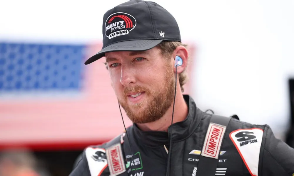 Cleetus McFarland Net Worth featured in a photo of race car driver and YouTuber Garrett Mitchell wearing a black racing suit and Tommy's Express cap outdoors. 