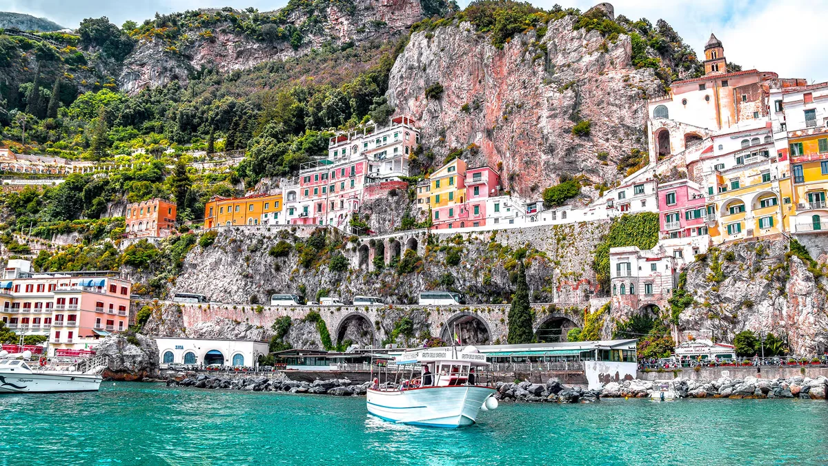 Colorful, historic buildings built into steep rocky cliffs along the turquoise waters of the Amalfi Coast with boats anchored in the foreground.