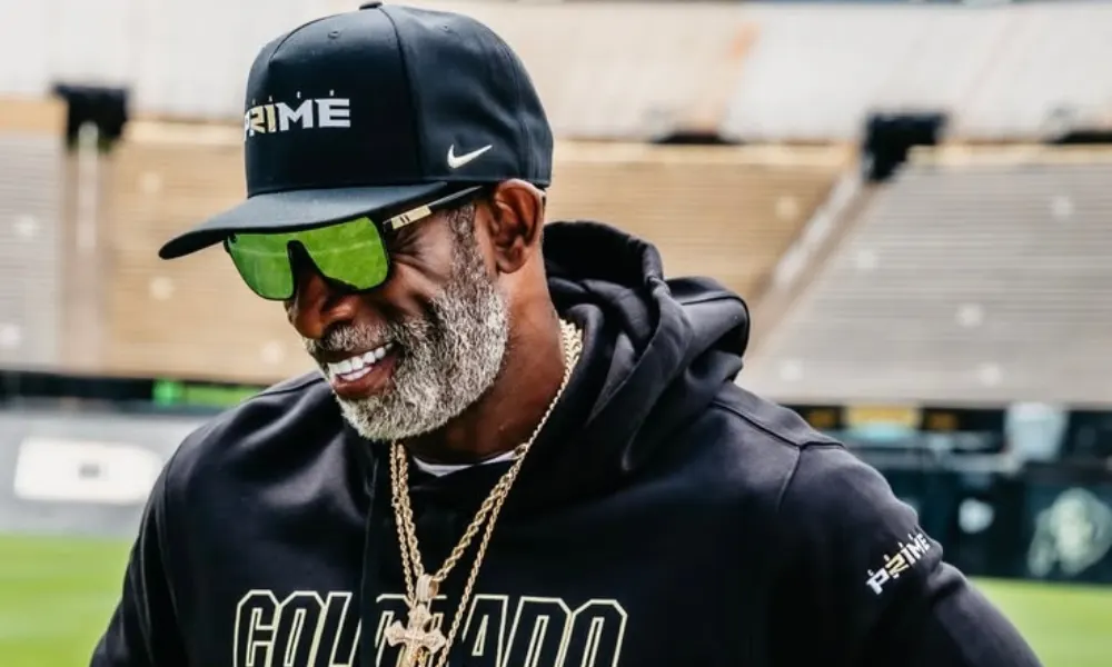  Deion Sanders in Colorado Buffaloes gear smiling at a football stadium. 