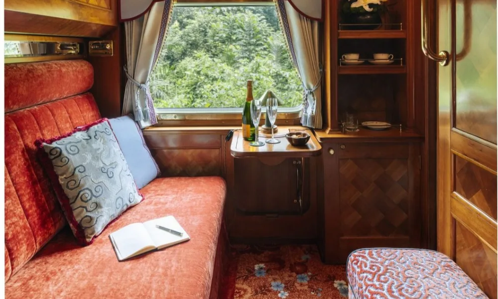 Elegant Eastern & Oriental Express train cabin with a velvet sofa, open journal, and champagne setup by a window showing lush green scenery, perfect for luxury train journeys.