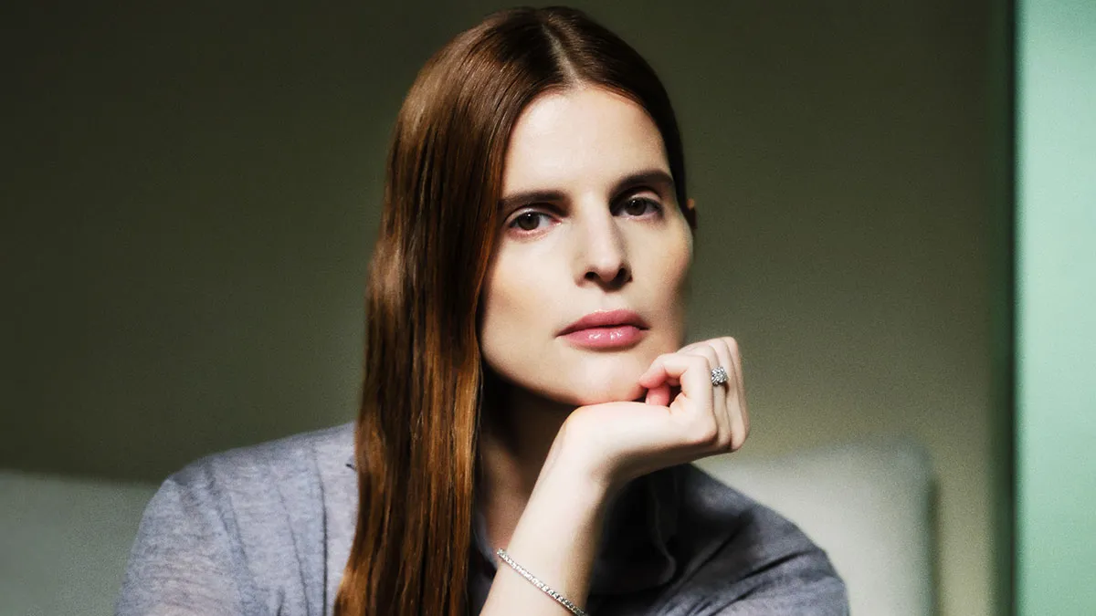 Close-up portrait of a woman with long straight brown hair resting her chin on her hand, wearing a gray shirt and silver jewelry.