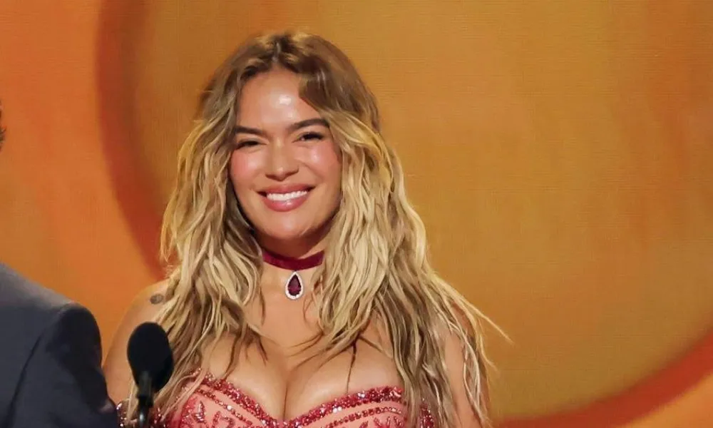 Karol g net worth highlighted in an image of Karol G smiling on stage in an embellished red sheer gown with a matching choker necklace. 