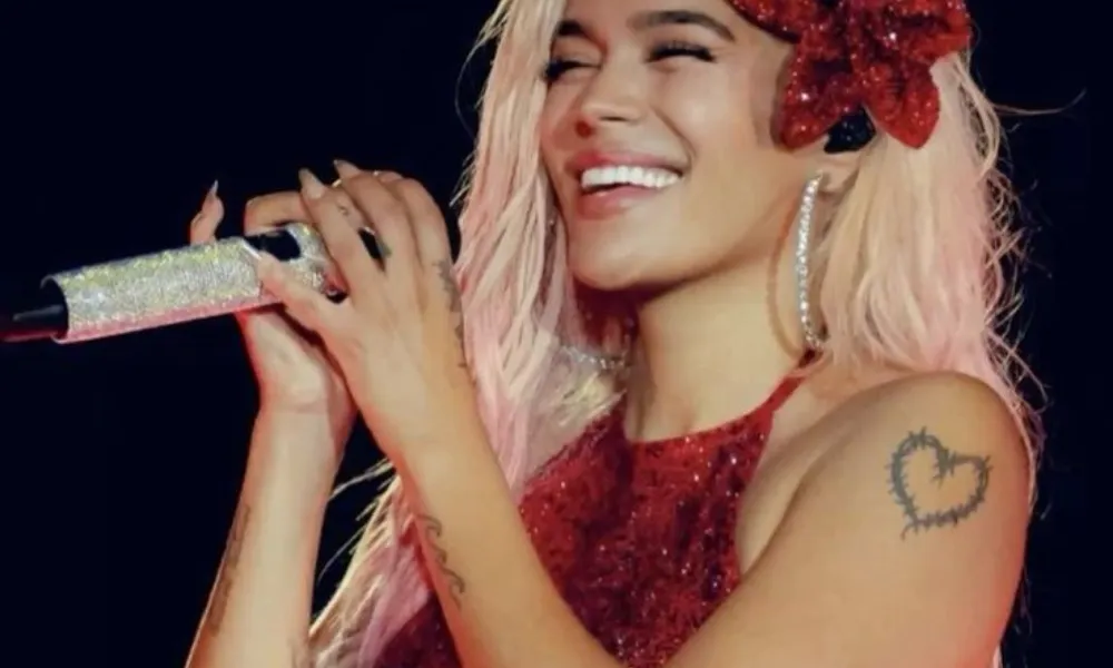 Karol g net worth featured in a image of Karol G singing on stage in a glittering red sequined dress with blonde hair and a red flower hairpiece. 