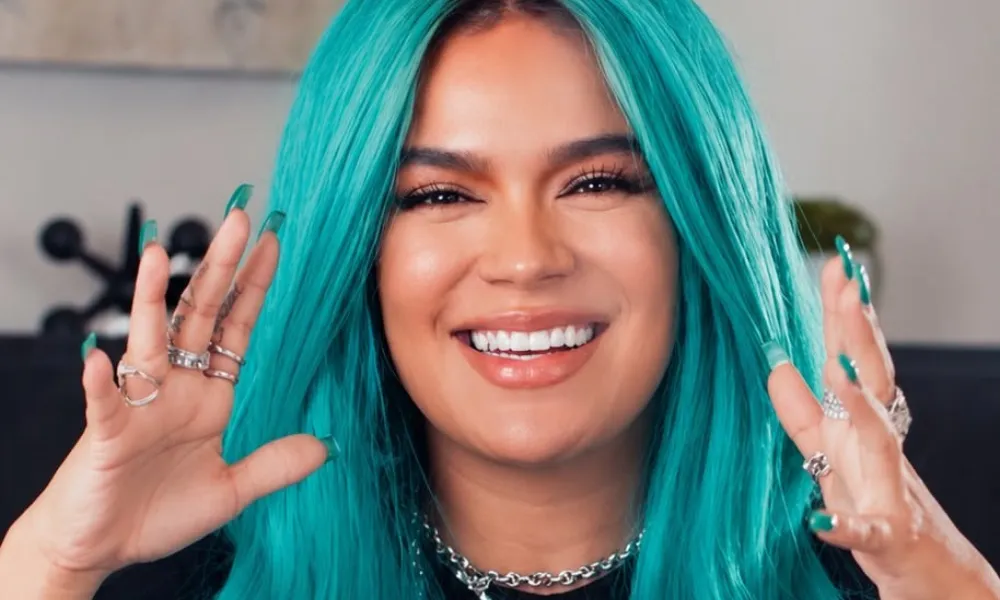 Karol G net worth highlighted in a portrait of singer Karol G smiling with vibrant turquoise hair, stacked rings, and a silver chain necklace. 