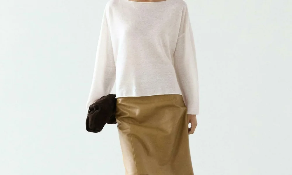 A studio shot of a model wearing an elegant lightweight white boatneck long-sleeve sweater paired with a camel beige leather midi skirt and holding a dark brown clutch, demonstrating sleek sophistication from old money fashion brands. 