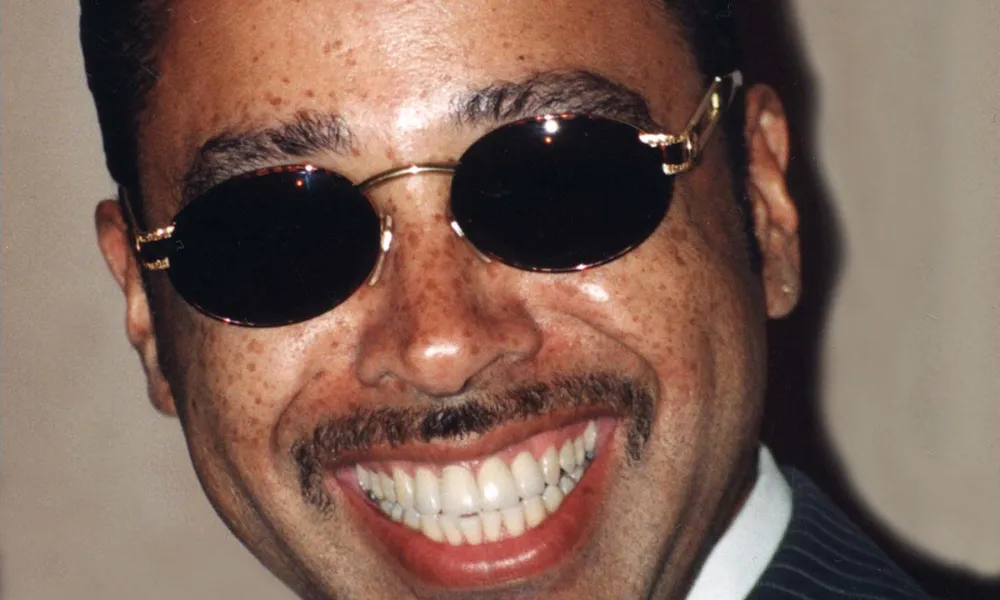Morris Day Net Worth featured in a portrait of the funk singer smiling in a pinstriped suit and dark oval sunglasses.