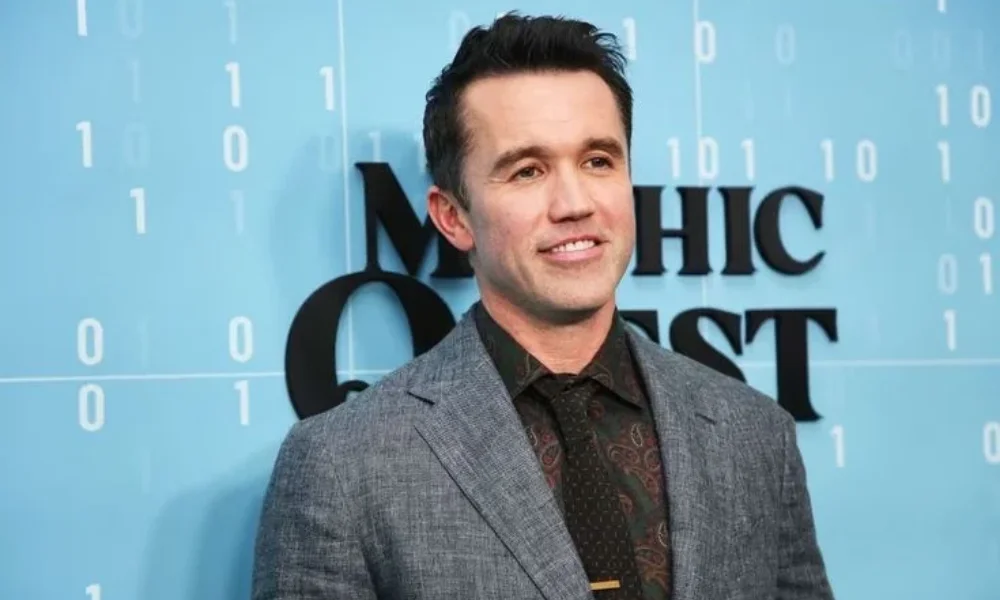Actor and producer Rob McElhenney posing at a red carpet premiere event for Mythic Quest against a blue backdrop, highlighting his Hollywood success and estimated Rob McElhenney Net Worth