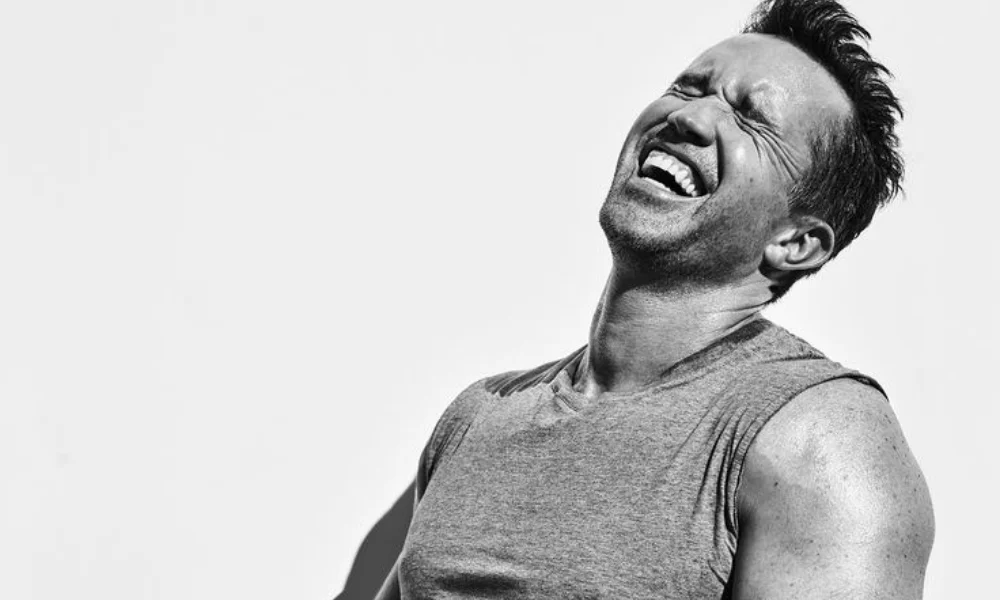 A black and white portrait of actor and producer Rob McElhenney laughing in a sleeveless shirt, reflecting his successful career in entertainment and estimated Rob McElhenney Net Worth