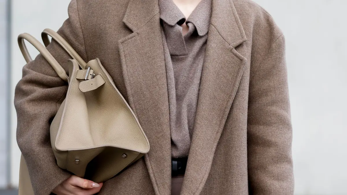 Close-up view of a person wearing a taupe wool coat over a matching collared sweater, carrying a structured beige leather handbag under their arm.