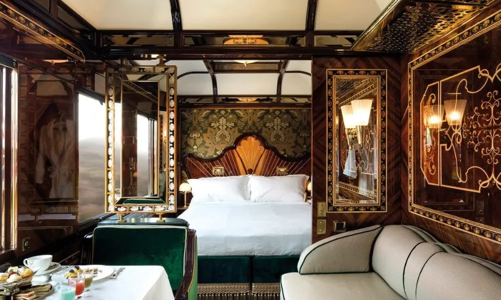 Luxurious Venice Simplon-Orient-Express cabin featuring a double bed, plush seating, elegant wood paneling, and a breakfast table set for luxury train journeys.
