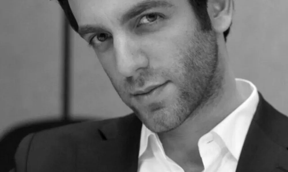 B.J. Novak Net Worth highlighted in a black-and-white portrait of B.J. Novak wearing a dark suit jacket over an open-collar white dress shirt. 