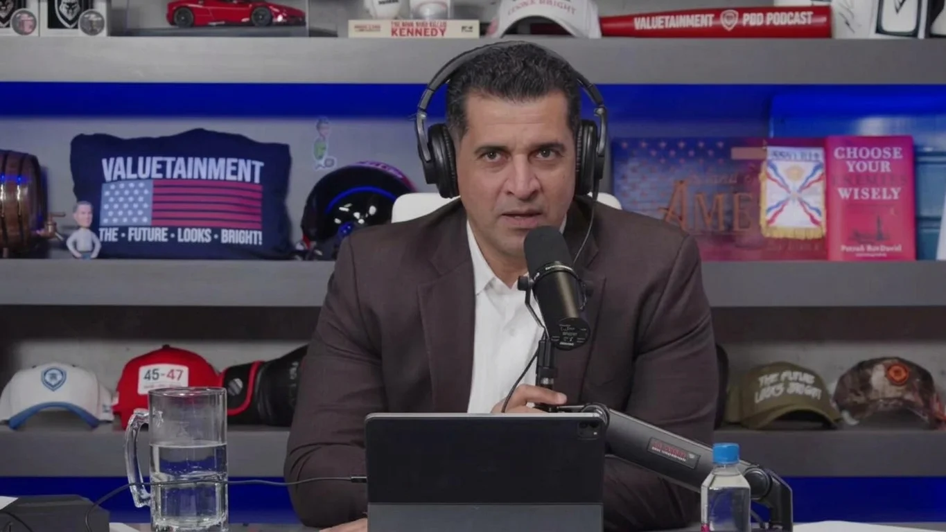 Patrick Bet-David sitting at a studio desk wearing headphones in front of a podcast microphone, backed by shelves filled with books and Valuetainment memorabilia.