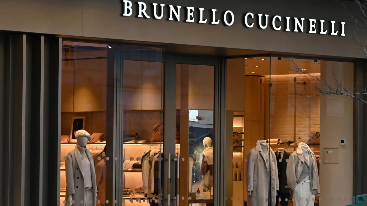 Storefront of a luxury Brunello Cucinelli boutique featuring glass display windows with mannequins in neutral-toned apparel.