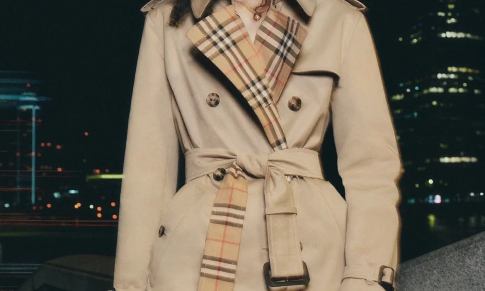 A close-up shot of a classic beige Burberry trench coat featuring its iconic plaid check lining on the lapels and belt, tied at the waist against a dark night cityscape, perfect for showcasing premier old money fashion brands. 