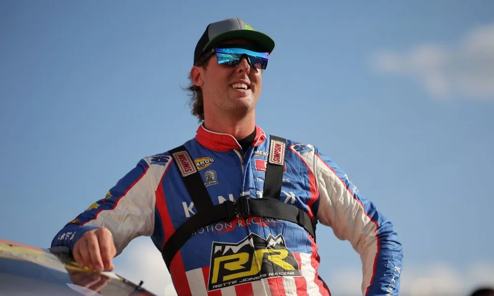 Cleetus McFarland Net Worth featured in a photo of YouTuber and race car driver Garrett Mitchell wearing a race suit and sunglasses leaning against his car. 