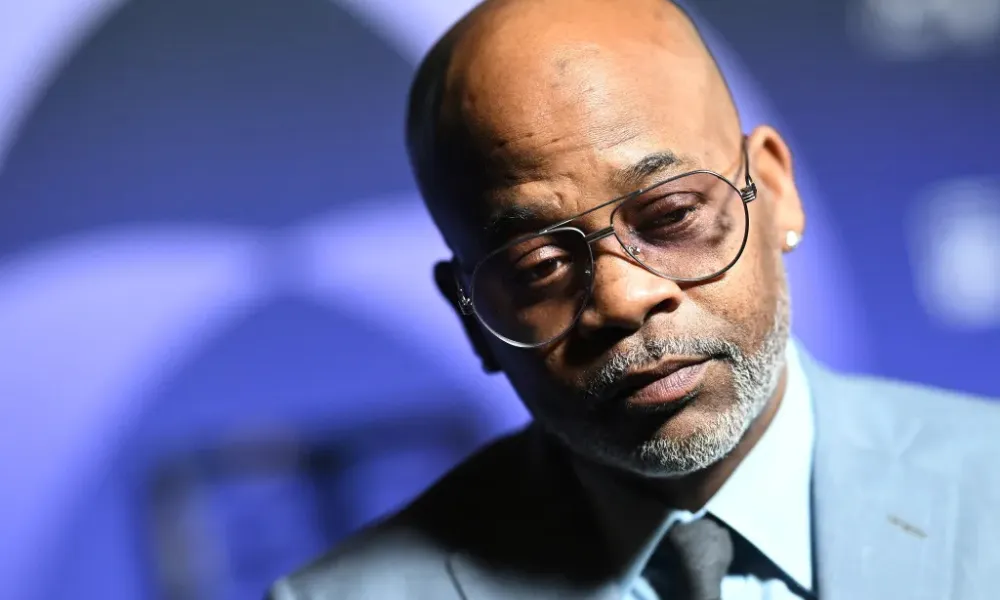 dame dash net worth highlighted in a close-up portrait of Damon Dash wearing wire-rimmed glasses, a light blue suit jacket, and a tie against a purple backdrop. 