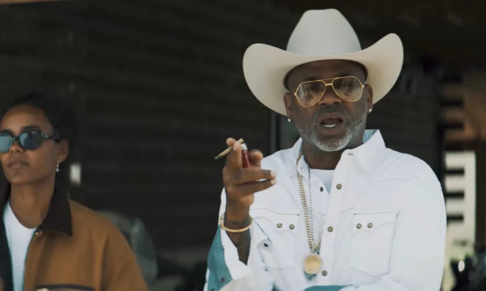 dame dash net worth highlighted in an image of Damon Dash wearing a white cowboy hat, sunglasses, and a gold chain while holding a cigar. 