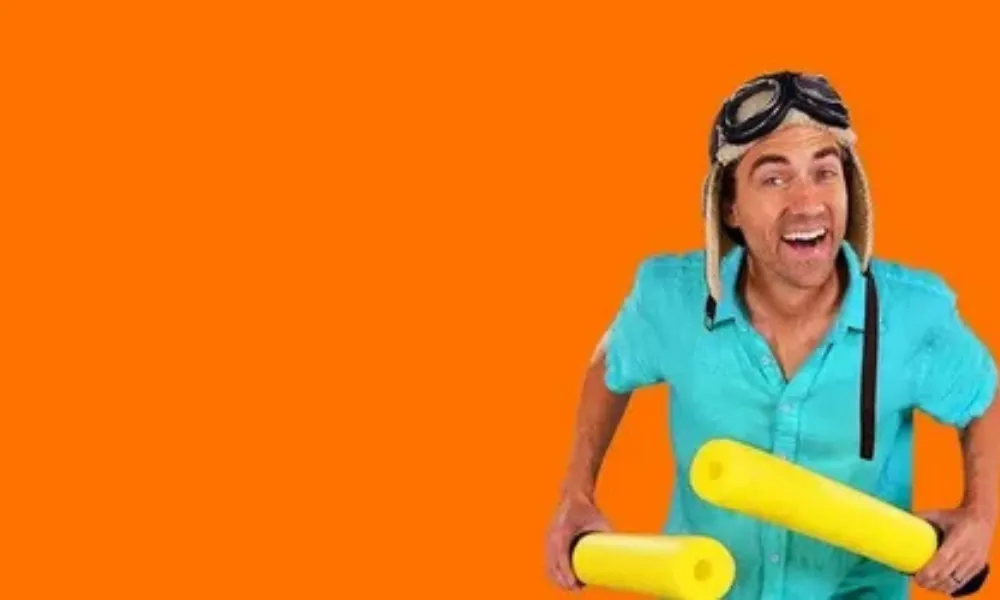 Danny Go Net Worth featured in a photo of children's host Danny Go holding yellow foam sticks in front of a bright orange background.