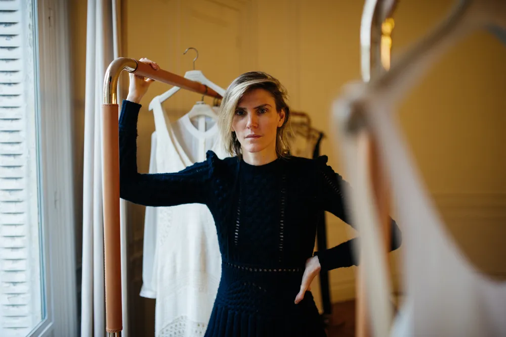 Woman standing beside a clothing rack with white dresses, holding the top bar while wearing a black long-sleeved dress.