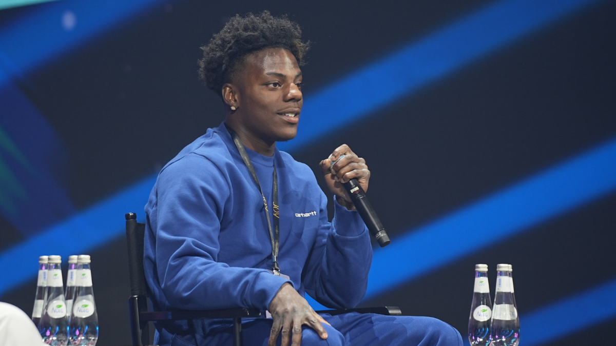 Streamer IShowSpeed speaking into a microphone on stage while wearing a blue Carhartt sweatshirt.