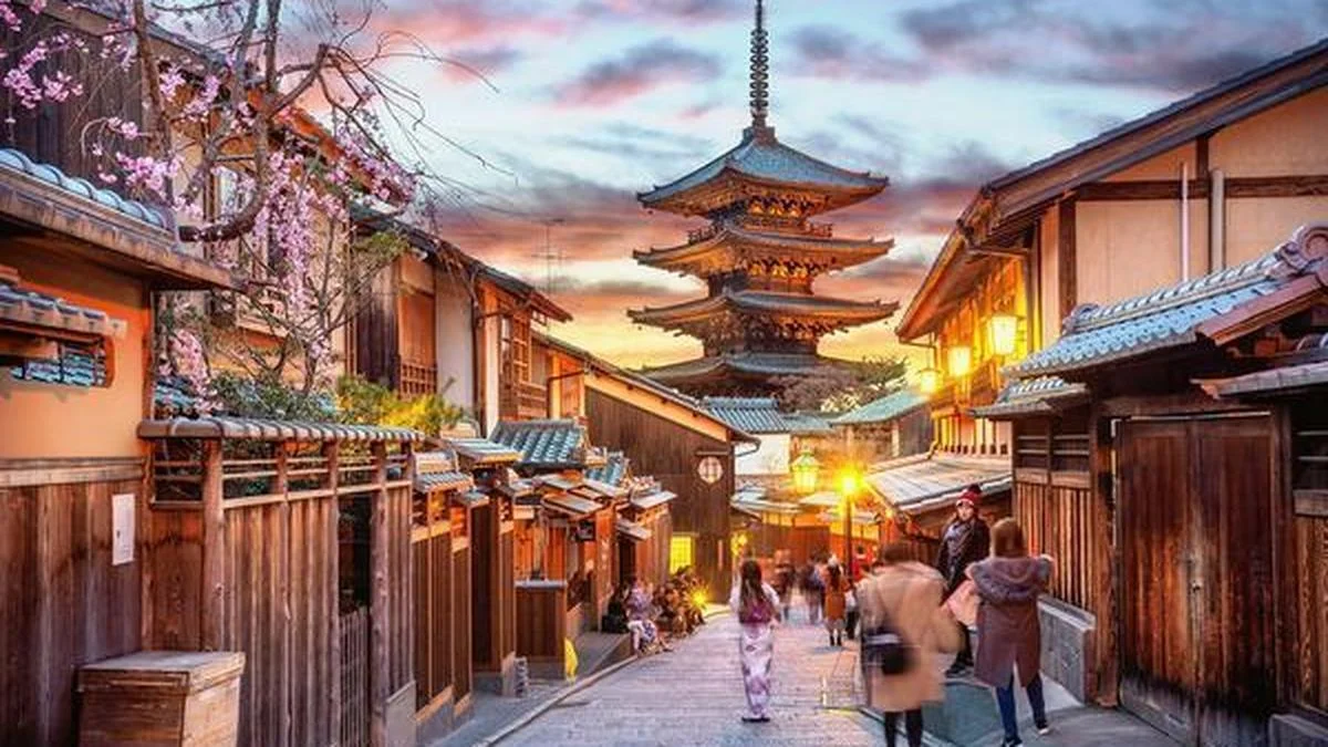 A historic street in Kyoto lined with traditional wooden buildings and blooming cherry blossoms, leading toward the five-story Yasaka Pagoda at sunset.