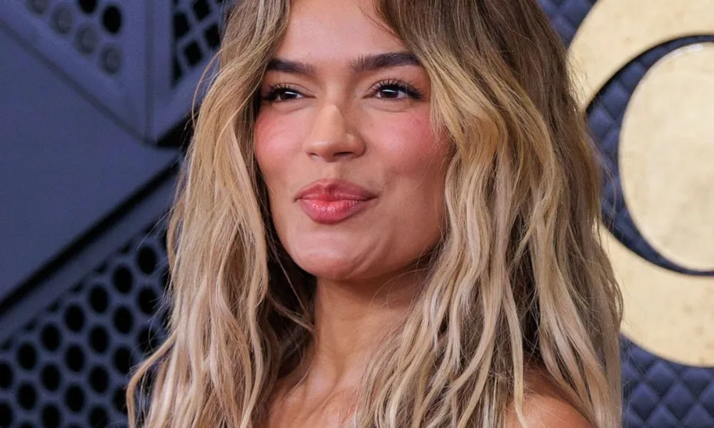 Karol g net worth highlighted in a close-up photo of Karol G with blonde wavy hair posing on a red carpet in an off-the-shoulder blue lace dress. 