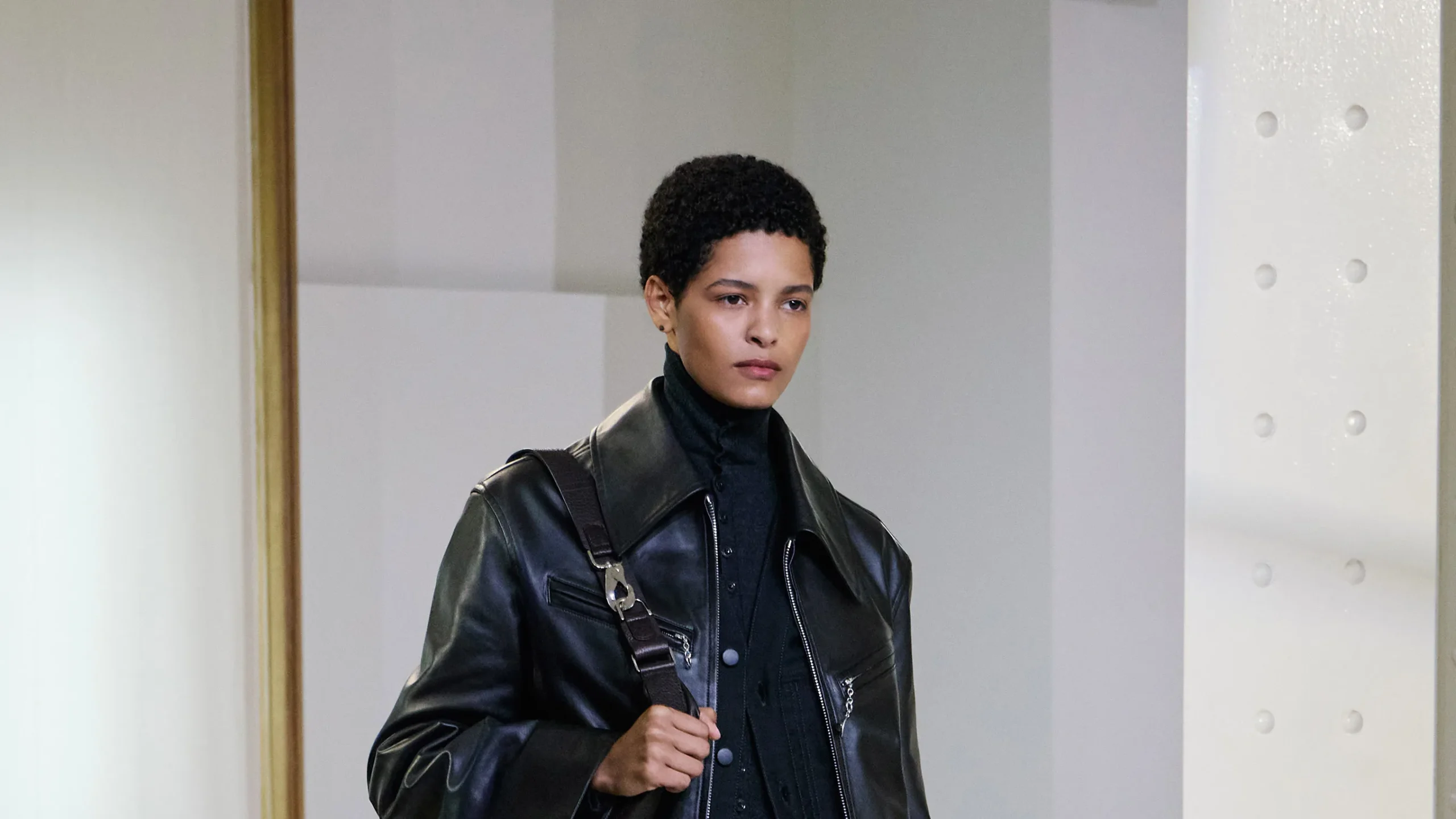 Model walking a runway wearing a black leather jacket over a dark turtleneck sweater with a shoulder bag strap.