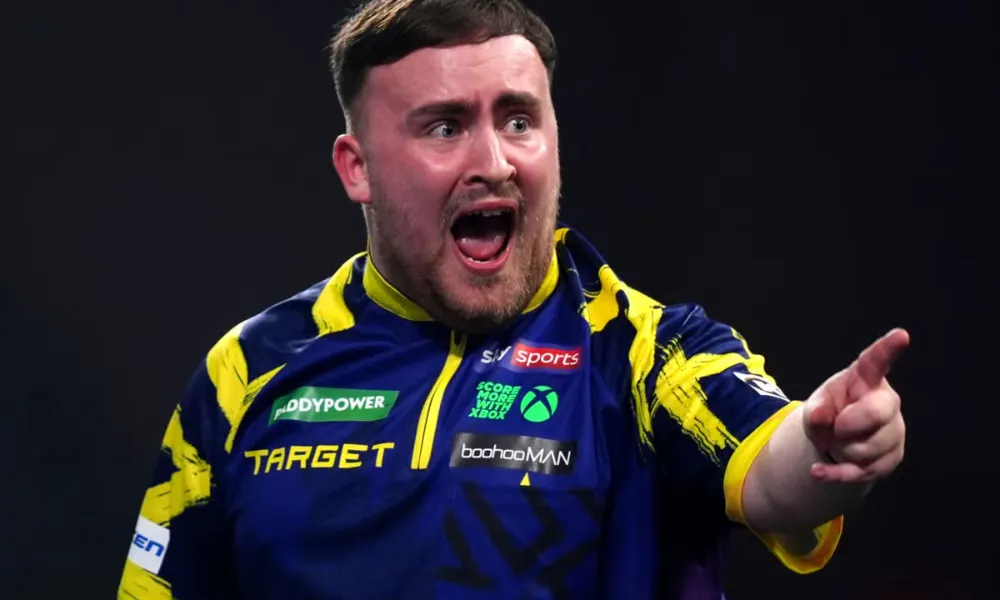  Luke Littler wears his blue and yellow dart shirt featuring major sponsor logos, visual context relevant to discussions surrounding Luke Littler net worth. 