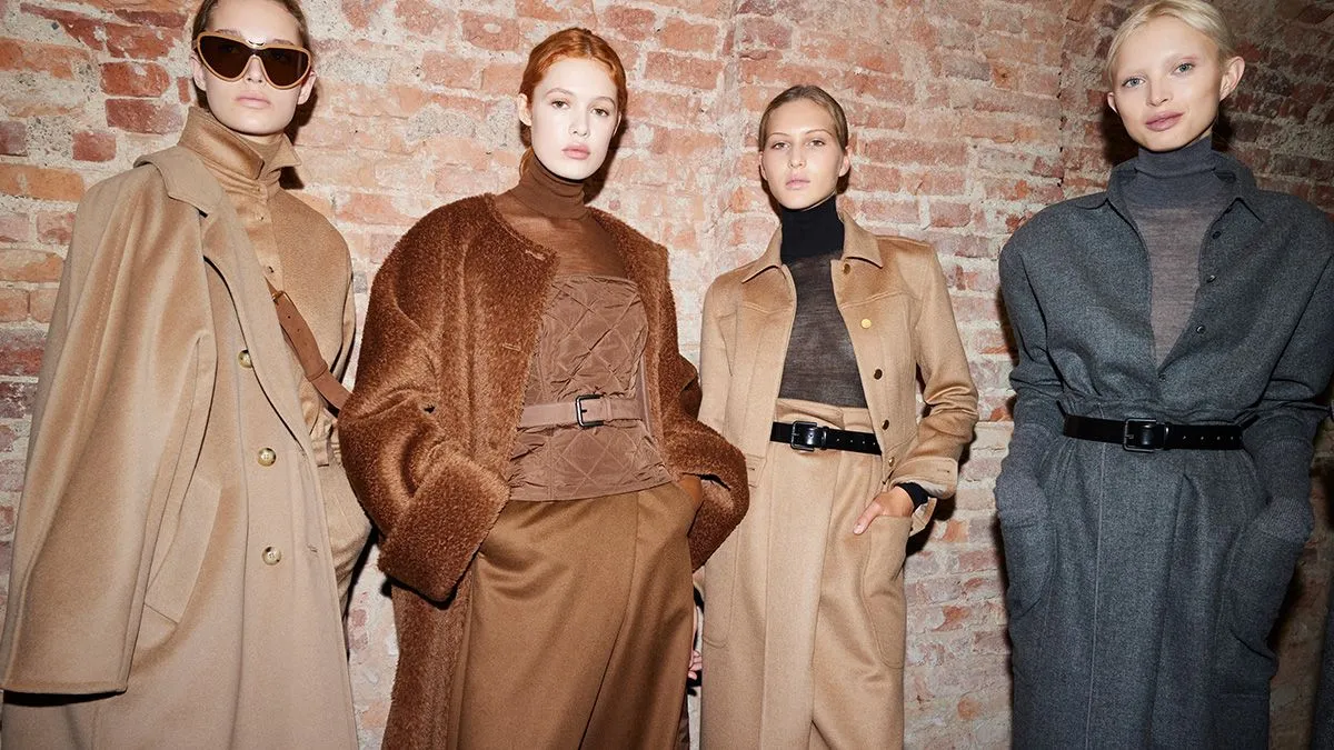 Four models standing in front of an exposed brick wall, wearing neutral-toned outerwear and tailored outfits.