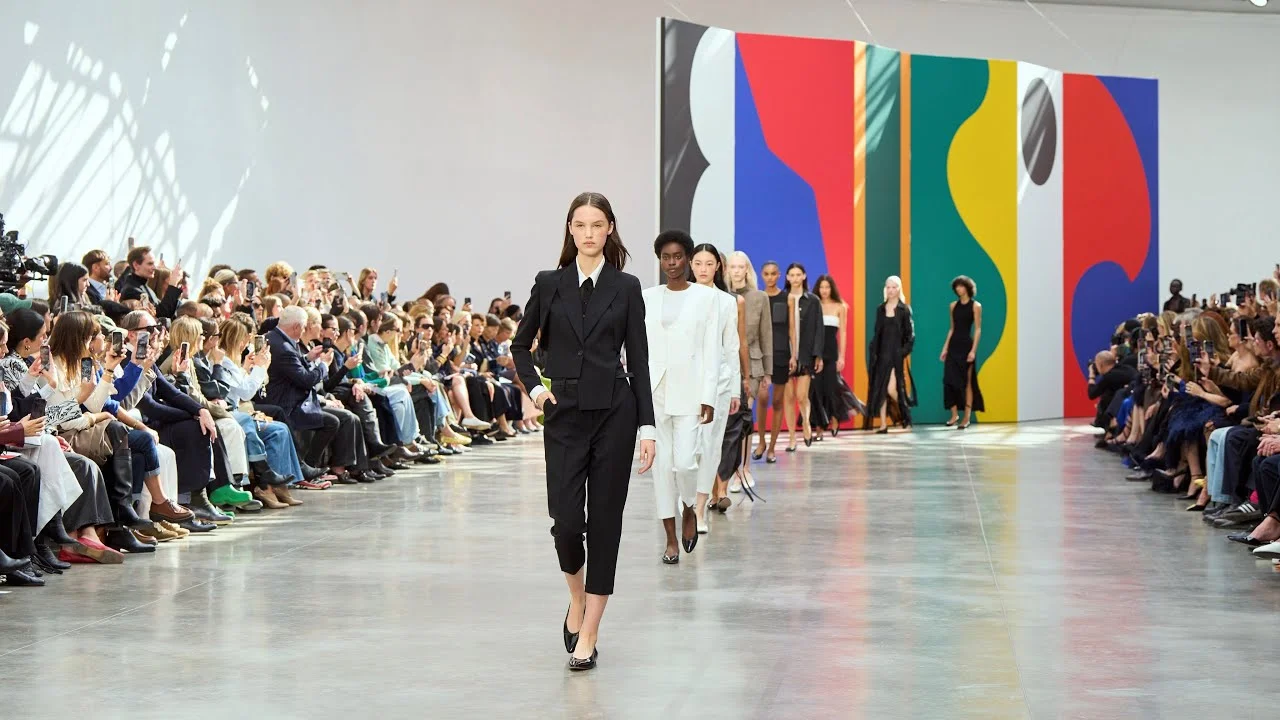 Models walking down a bright runway at a fashion show in front of a seated audience and a colorful geometric backdrop.