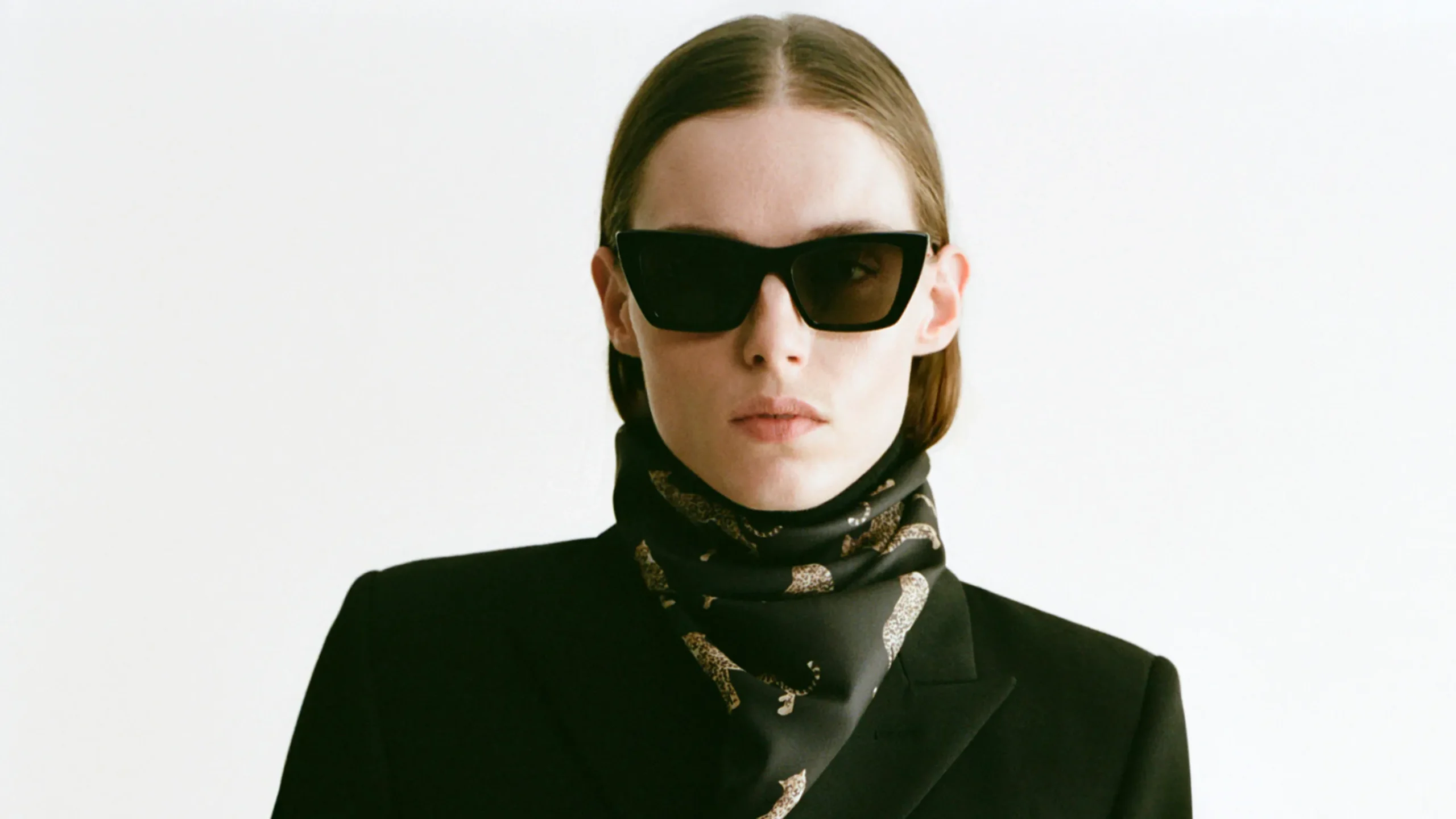 Close-up portrait of a woman wearing black sunglasses, a black blazer, and a dark neck scarf patterned with leopards.