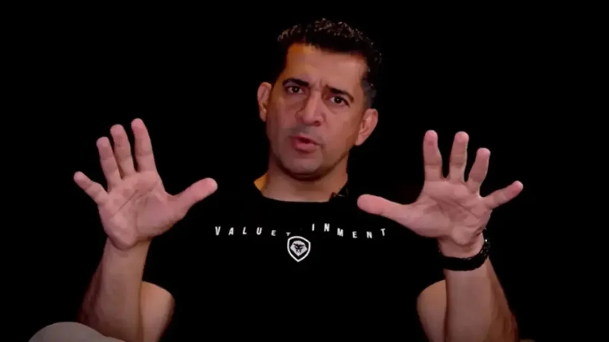 A medium shot of Patrick Bet-David raising both hands with open palms while wearing a black Valuetainment t-shirt against a solid black background.