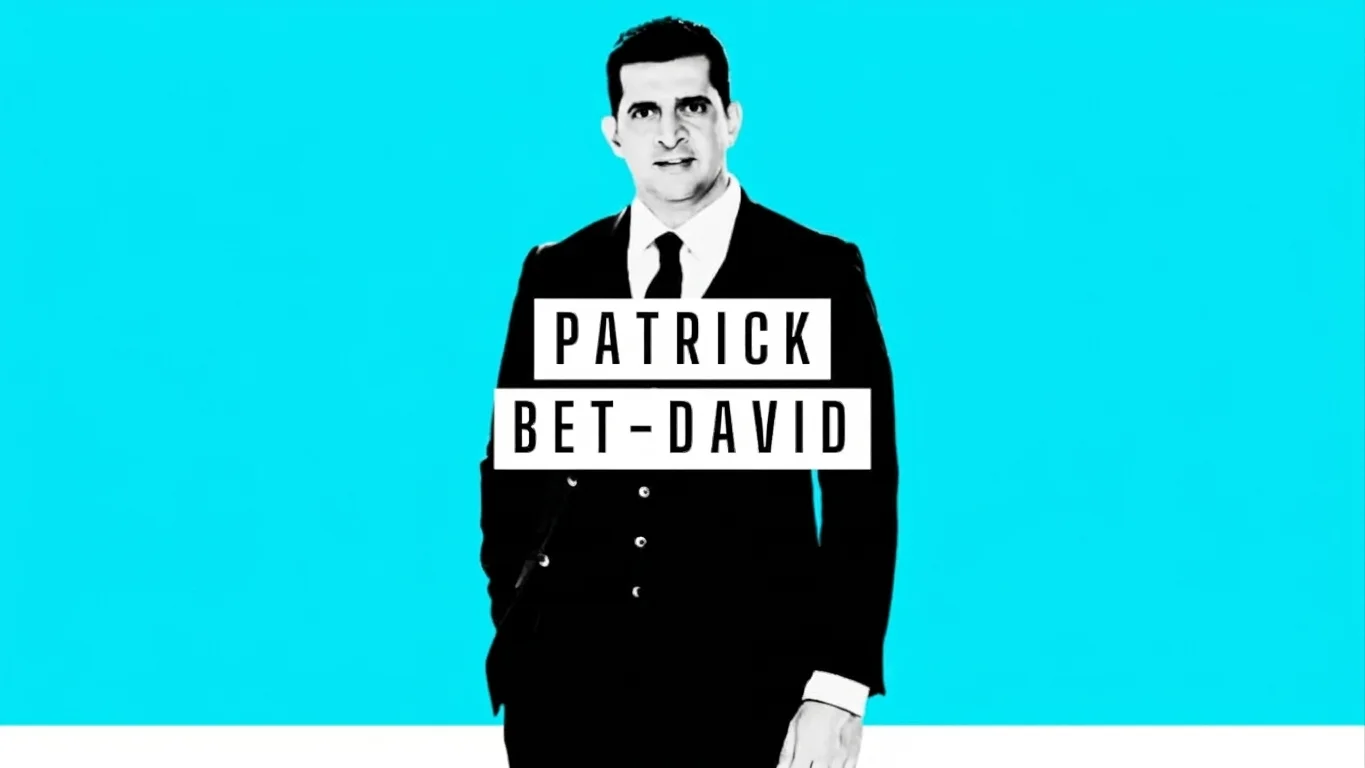 Patrick Bet-David seated in front of a podcast microphone set against a black-and-white comic-style backdrop of city skyscrapers featuring his name in large graphic letters.