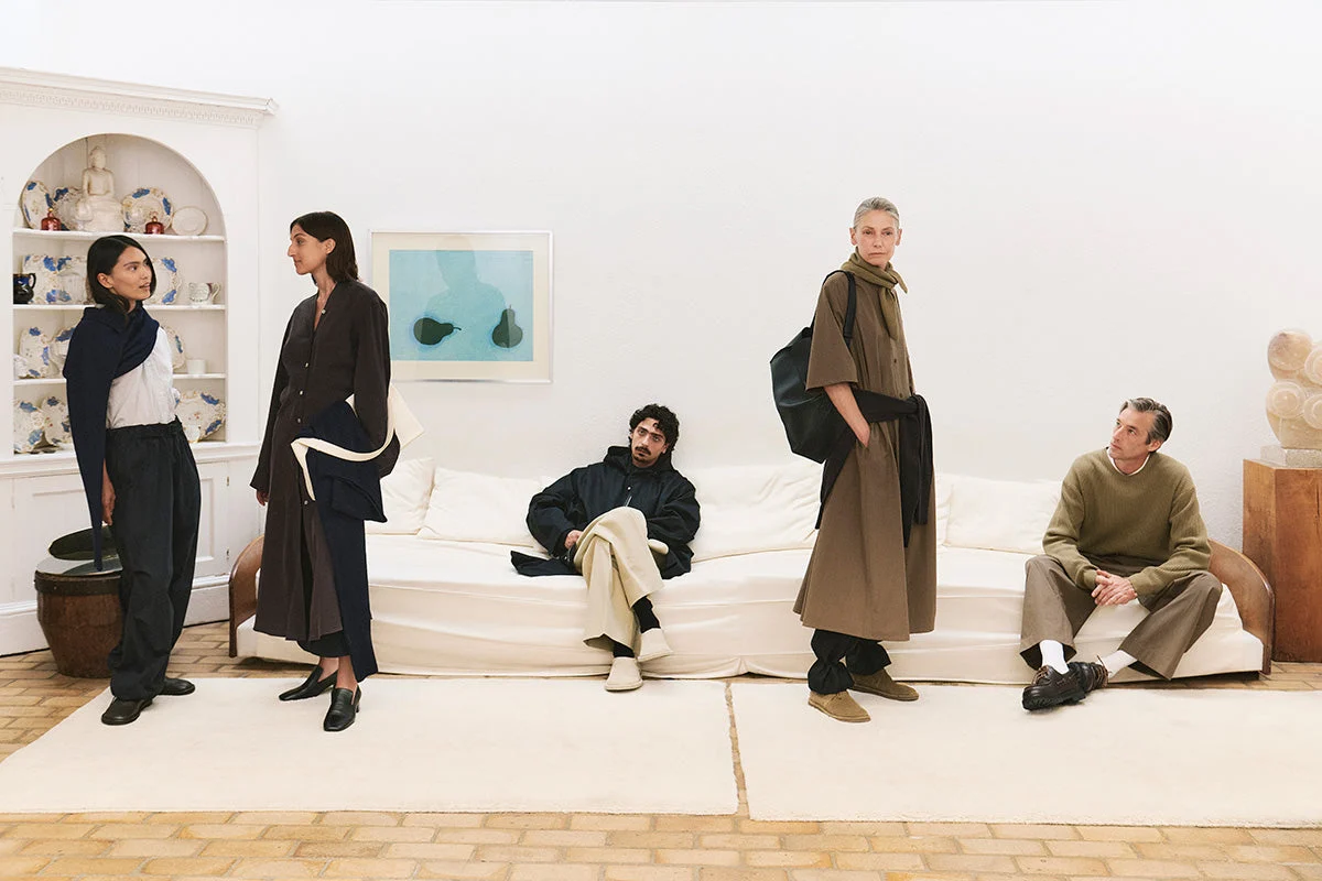Group of models in relaxed, minimalist earth-toned clothing posing in a modern white interior room with a sofa.