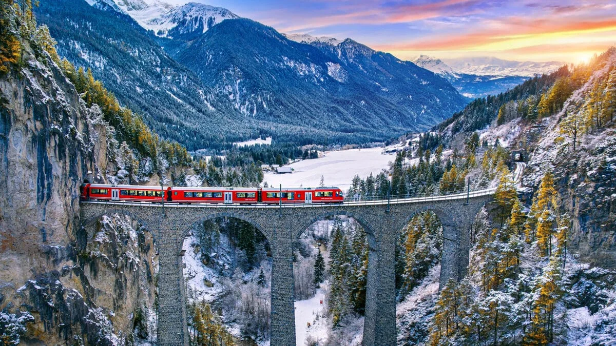 Red train crossing the Landwasser Viaduct surrounded by snow-dusted mountains and pine forests in Switzerland.